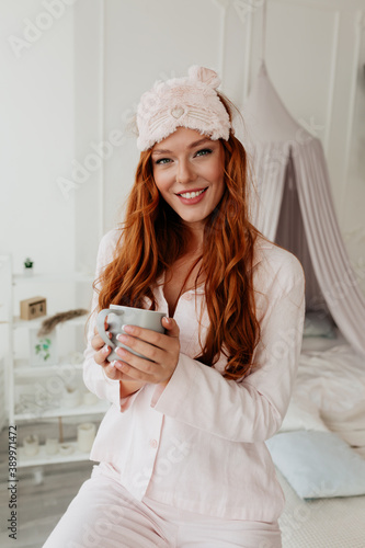 Smiling adorable lady with red hair wearing pajams holding coffee looking at camera on background of her house photo