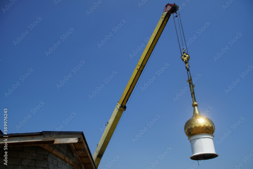 A crane lifted the bright golden dome of the Christian church for installation and assembly on the roof of the temple under construction. Blue sky. A crane lifted the bright golden dome of the Christian church for installation and assembly on the roof of the temple under construction. Blue sky.