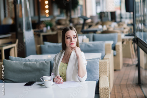Young woman in white jacket working in cafe with papers, drinking coffee
