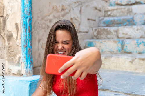 attractive girl dressed in red dress taking a selfie in the street on a sunny day and smiling © Supermelon