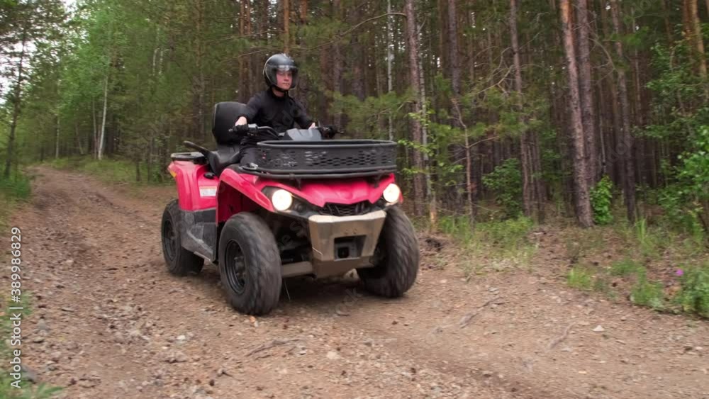 Handheld shot of man in helmet driving red quad bike downhill along dirt road in forest