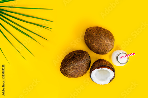 Top view coconut water drink, milk in a bottle with straw, and fresh coconuts with a green palm leaf on the yellow background. Summer exotic refreshment. Natural plant based food. Flatlay. Copy space.