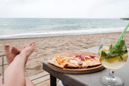Fototapeta Naklejka Na Ścianę i Meble -  Young Caucasian woman lying at the beach eating pizza and drinking cocktail. Legs, sea in background. Careless holidays at all inclusive resort. Luxury recreational vacation concept at Mediterranean
