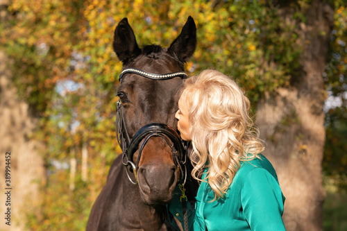 Young blond woman in green dress, kissing a brown horse. In the forest at autumn in golden color. love horses