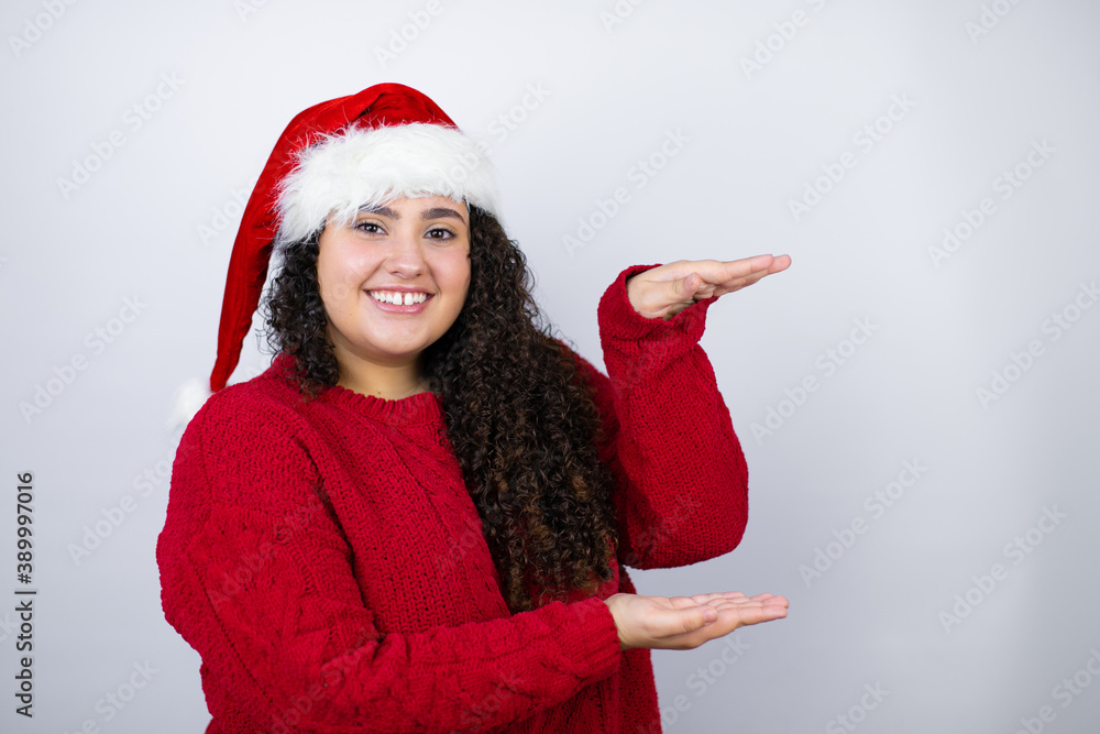 Young beautiful woman wearing a Santa hat over white background gesturing with hands showing big and large size sign, measure symbol. Smiling . Measuring concept. Young beautiful woman wearing a Santa hat over white background gesturing with hands showing big and large size sign, measure symbol. Smiling . Measuring concept.