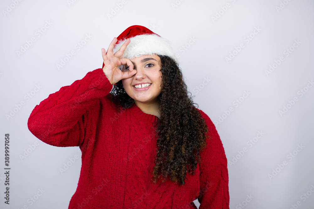 Young beautiful woman wearing a Santa hat over white background doing ok gesture shocked with smiling face, eye looking through fingers Young beautiful woman wearing a Santa hat over white background doing ok gesture shocked with smiling face, eye looking through fingers