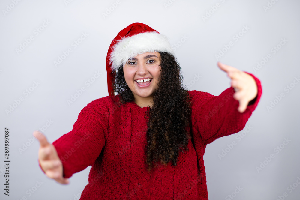 Young beautiful woman wearing a Santa hat over white background looking at the camera smiling with open arms for hug Young beautiful woman wearing a Santa hat over white background looking at the camera smiling with open arms for hug