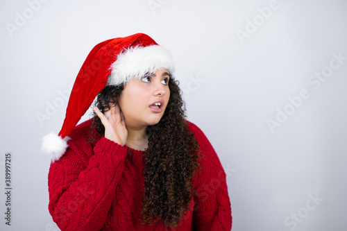 Young beautiful woman wearing a Santa hat over white background surprised with hand over ear listening an hearing to rumor or gossip