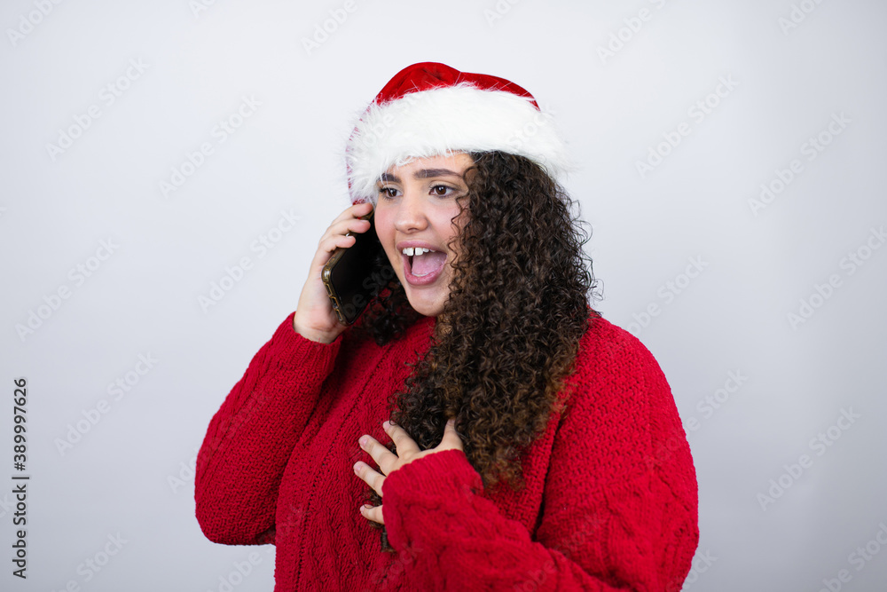Young beautiful woman wearing a Santa hat over white background talking on the phone with a worried expression Young beautiful woman wearing a Santa hat over white background talking on the phone with a worried expression