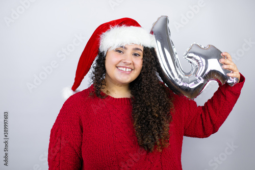 Young beautiful woman wearing a Santa hat over white background smiling holding a number four balloon. Celebration concept