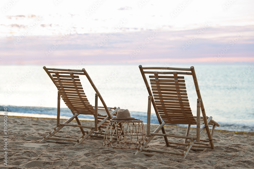 Wooden deck chairs and wicker table on sandy beach at sunset. Summer vacation