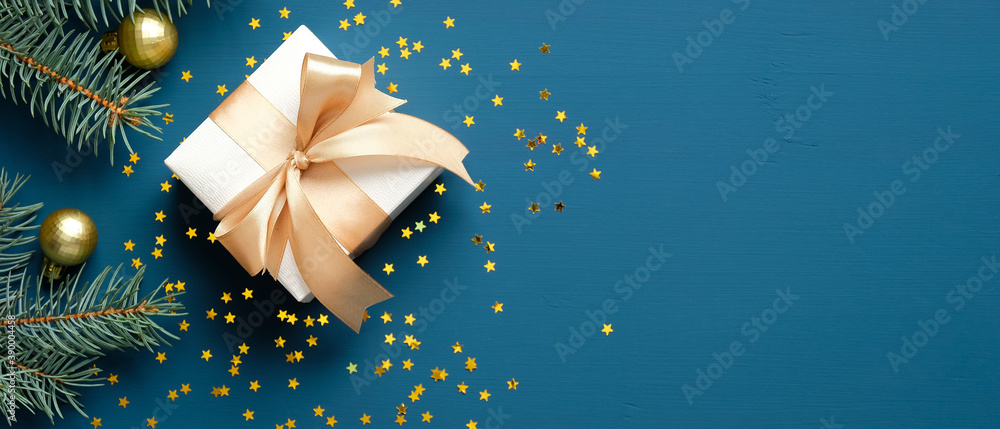Christmas gift box with golden ribbon bow, fir branches, confetti on dark blue background. Xmas banner design, header mockup. Flat lay, top view, copy space. - obrazy, fototapety, plakaty 