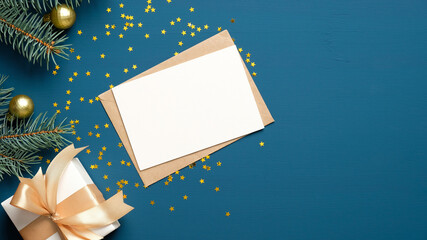 Christmas letter envelope with blank paper card, white gift box with golden ribbon bow, fir branches decorated balls on dark blue background. Flat lay, top view. Letter to Santa Claus concept.