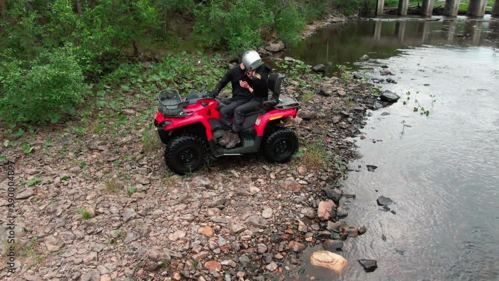 Aerial tracking shot of happy couple in helmets sitting on red quad bike by the river and kissing Aerial tracking shot of happy couple in helmets sitting on red quad bike by the river and kissing