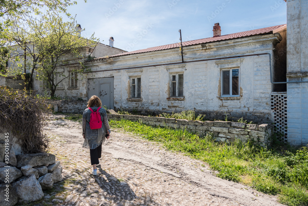 A woman with a backpack on the street with a dirt road and houses built in the 19th century in the city of Kerch in the Crimea