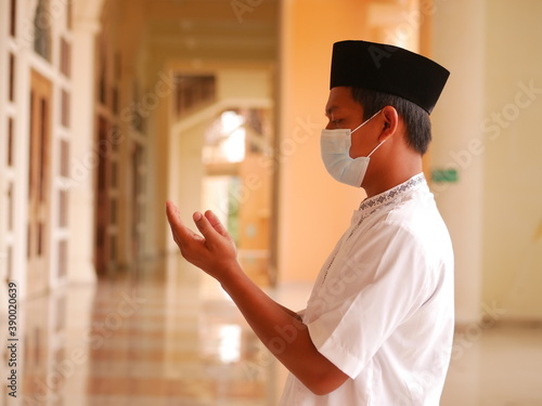 Indonesian muslim young man wearing medical face mask standing while raised hands and praying. Indonesiam moslem man praying with the prayer beads in his hand. photo