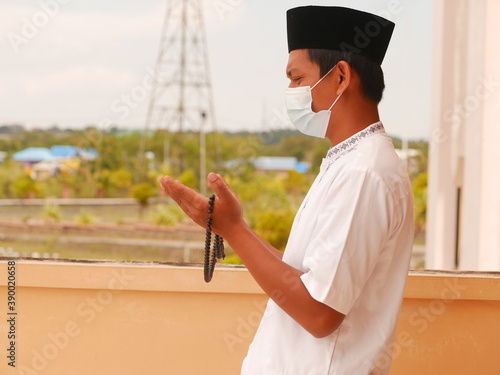 Indonesian muslim young man wearing medical face mask standing while raised hands and praying. Indonesiam moslem man praying with the prayer beads in his hand.