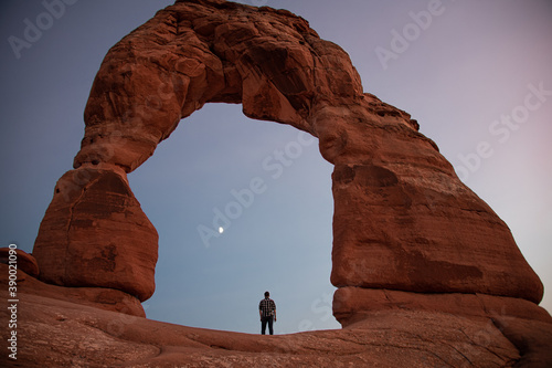 Person standing under the Delicate Arch in Arches National Park during sunset. Person looks tiny compared to the large famous rock formation.