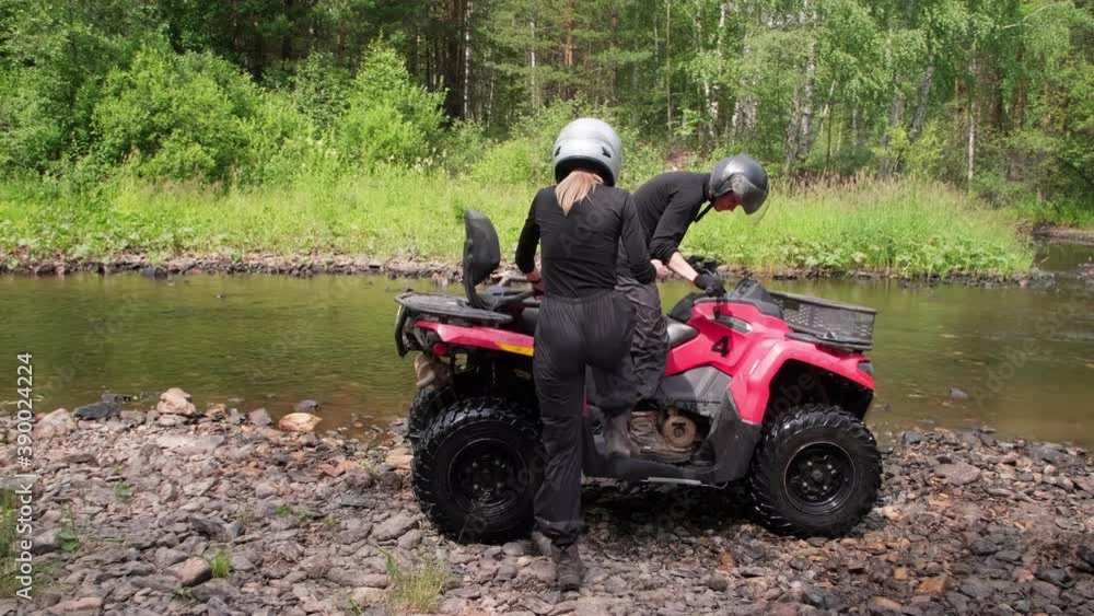 Tracking shot of man and woman in helmets getting on red quad bike and driving across shallow river in forest Tracking shot of man and woman in helmets getting on red quad bike and driving across shallow river in forest