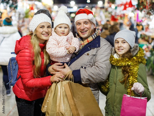 Portrait of cheerful man and woman and their happy daughters posing with paper bags near counters at Christmas fair