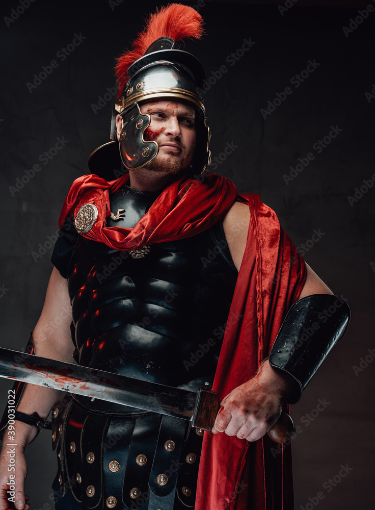 Naklejka premium Strong and warlike legionary with blood on his face in black armour with red cape poses holding a sword and looking away.