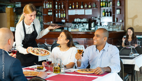 Hospitable young waitress bringing delicious pizzas to guests of cozy italian pizzeria