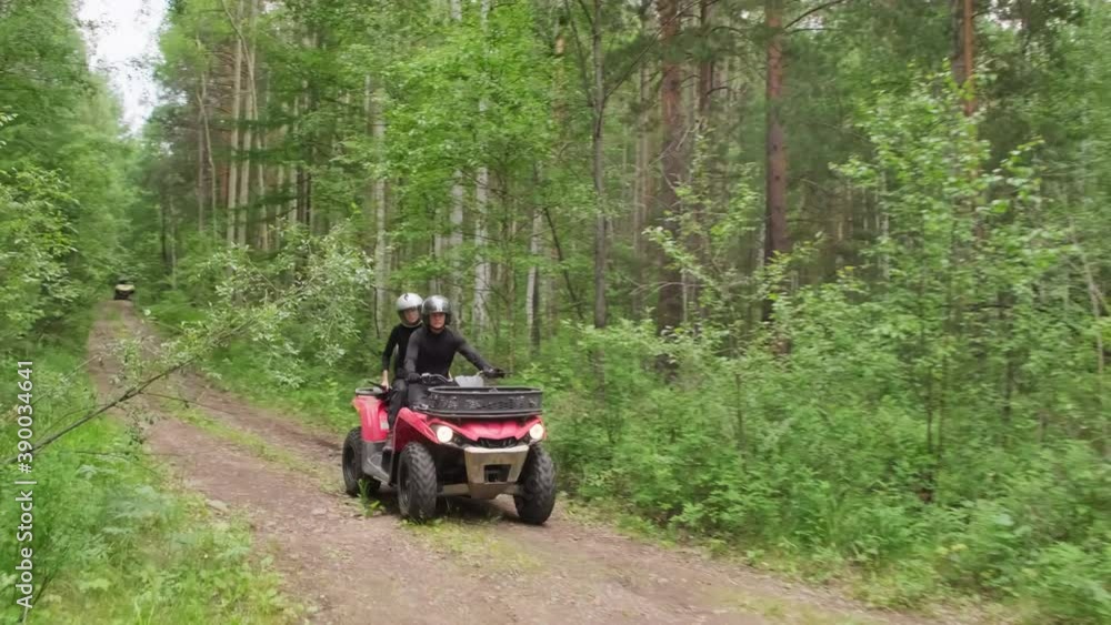 Handheld tracking shot of couple in helmets driving red quad bike along narrow forest road in rough terrain Handheld tracking shot of couple in helmets driving red quad bike along narrow forest road in rough terrain