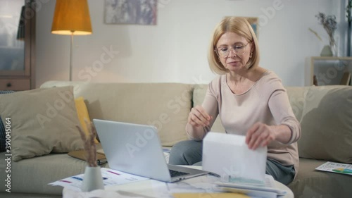 Middle aged caucasian woman in glasses working from home checking documents and statements unfolding letter with good news surprising and smiling