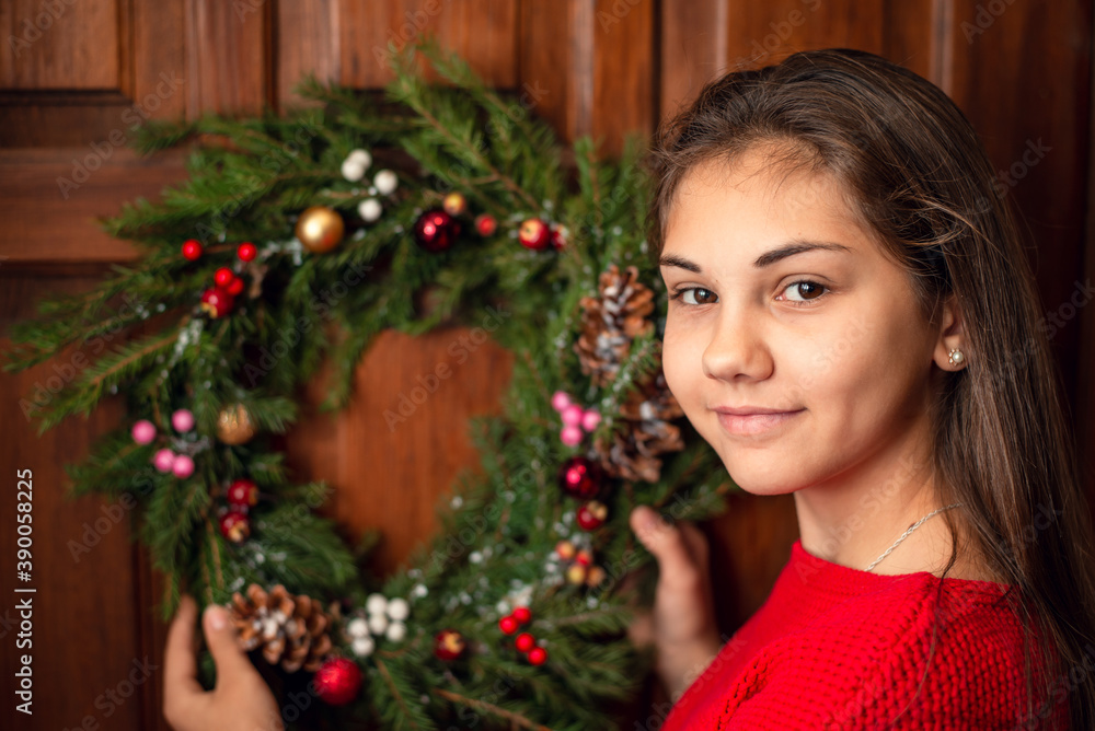 Young girl in red sweater hanging a Christmas wreath to the door of her home. Smiling person near the entrance of the house in winter time.