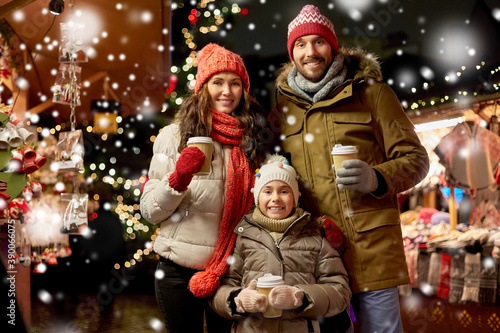 family, winter holidays and celebration concept - happy mother, father and little daughter with takeaway drinks at christmas market on town hall square in tallinn, estonia over snow
