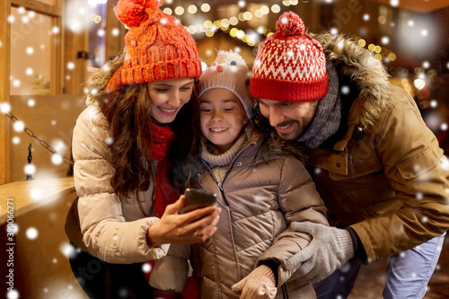 family, winter holidays and technology concept - happy mother, father and little daughter with smartphone at christmas market in evening over snow