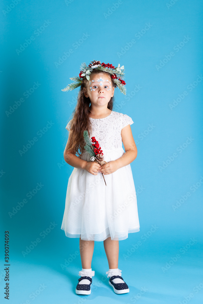 a full-length surprised girl in a white angel dress in a wreath holds a sprig of a Christmas tree isolated on a blue background. a full-length surprised girl in a white angel dress in a wreath holds a sprig of a Christmas tree isolated on a blue background.