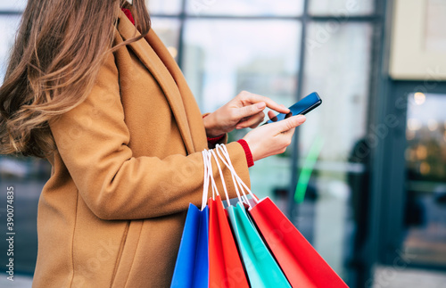 Beautiful trendy young woman with many colorful shopping bags in good mood with smart phone and credit card while walking in the mall during black friday