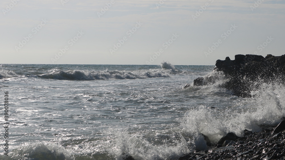 Fototapeta premium silvery waves and foamy sparkling spray of the sea surf beat against the black rocks of the deserted coast in the sun glare