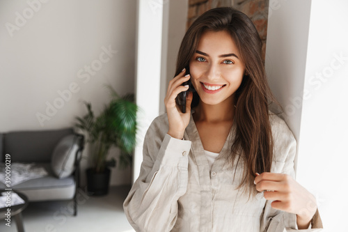 Young woman talking on phone near window. Girl calling someone from home and smiling