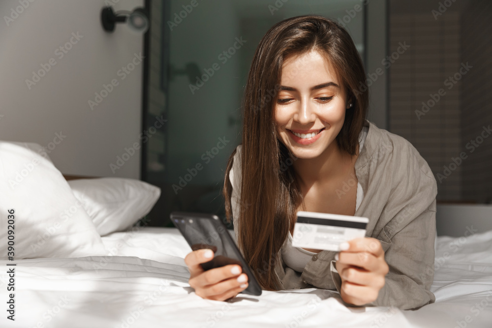 Young woman with smartphone and credit card lying in bed at home, shopping online