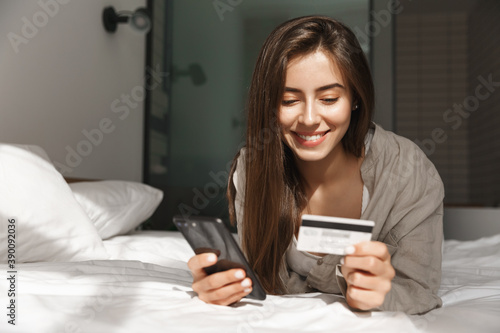 Young woman with smartphone and credit card lying in bed at home, shopping online