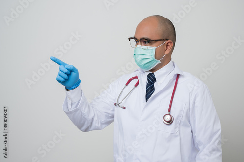 Asian bald Doctor in white coat  stethoscope  blue glove wearing face mask.pointing finger to beside camera isolated on white background.