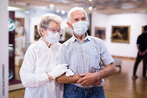 mature couple in mask protecting against covid examines paintings on display in hall of art museum photo