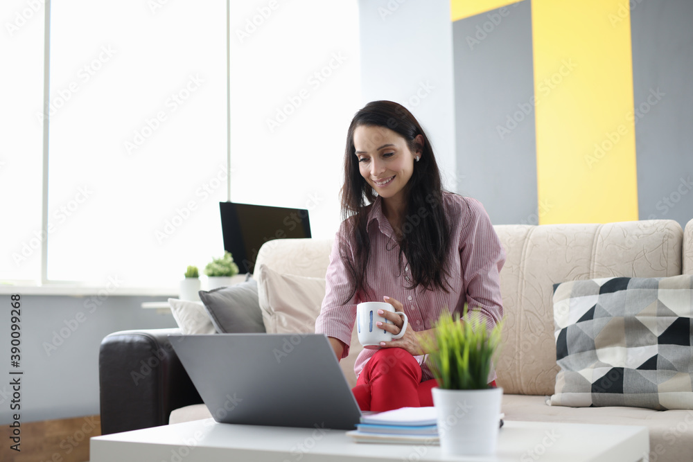 Woman is sitting on couch holding cup in her hands and working at laptop. Remote work at home for freelancers concept Woman is sitting on couch holding cup in her hands and working at laptop. Remote work at home for freelancers concept