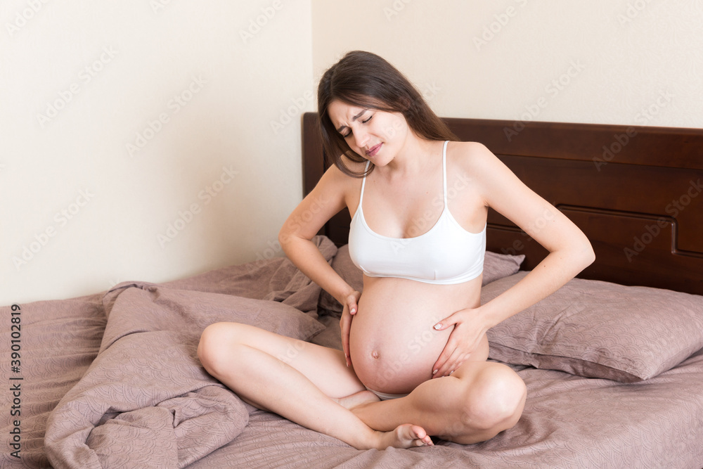 Young pretty pregnant woman, in pain, sitting on bed in bedroom, preparing to give a birth Young pretty pregnant woman, in pain, sitting on bed in bedroom, preparing to give a birth