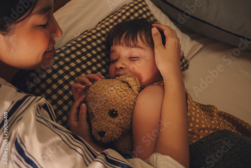 Mother watching her child sleep and hug teddy bear on a bed at night.