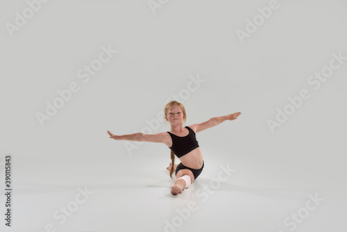 Full length shot of little redhead girl, professional gymnast looking away, doing splits, showing flexibility isolated over grey background