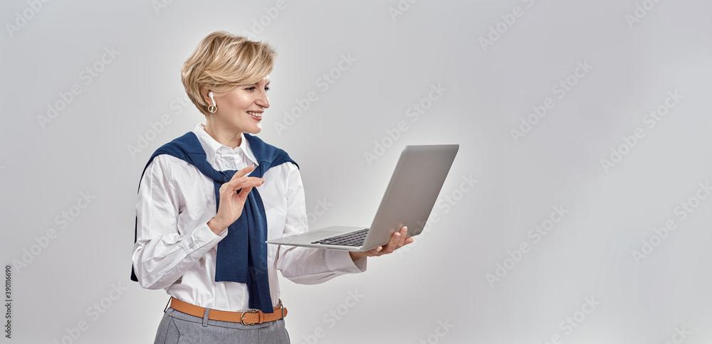 © Svitlana - Portrait of elegant middle aged caucasian woman wearing business attire holding laptop, while having online video call, standing isolated over grey background
