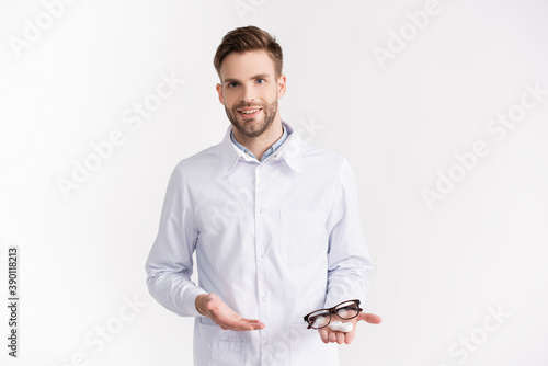 Front view of smiling doctor pointing with hand at eyeglasses and lenses container on palm isolated on white