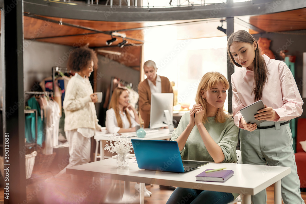 A young well-dressed woman holding a tablet sharing ideas with her young female coworker with their collaborateurs on a background