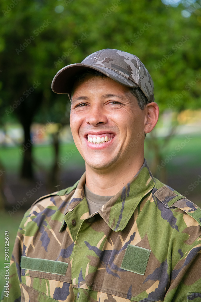 Cheerful man in military camouflage uniform standing in park, looking at camera and smiling. Green trees in background. Vertical shot. Military man or guard concept