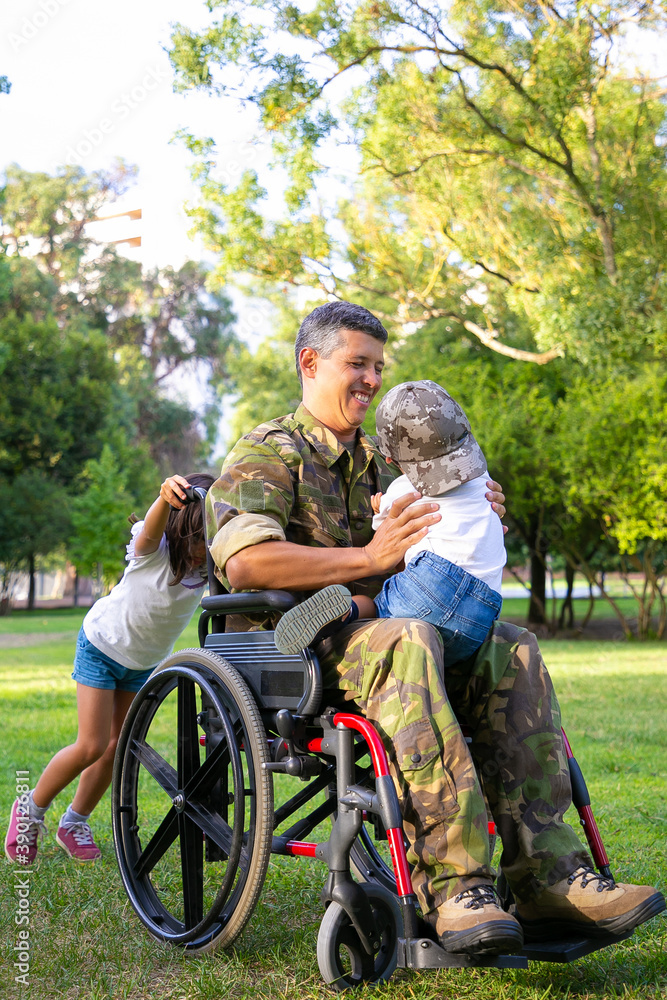 Joyful disabled military dad walking with two children in park. Girl pushing wheelchair handles, boy sitting on dads lap. Veteran of war or disability concept