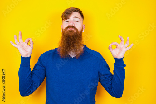 Young handsome red haired bearded man standing against yellow wall doing yoga, keeping eyes closed, holding fingers in mudra gesture. Meditation, religion and spiritual practices. photo