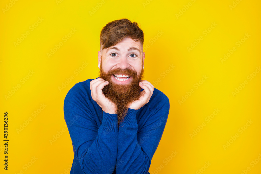 Dreamy Young handsome red haired bearded man standing against yellow wall keeps hands pressed together under chin, looks with happy expression, has toothy smile. Dreamy Young handsome red haired bearded man standing against yellow wall keeps hands pressed together under chin, looks with happy expression, has toothy smile.